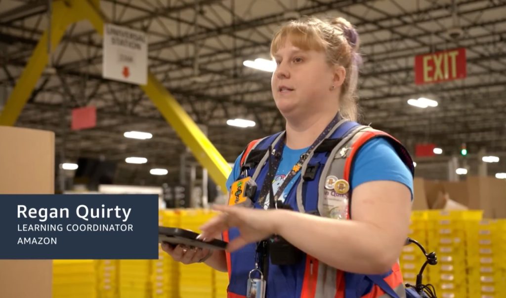 Amazonのフルフィルメントセンターで、青・赤・グレーのテックベストを着用したAmazon従業員が解説する様子。背後に黄色の棚と出口サインが見える。
